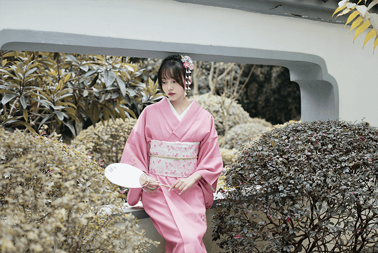 Pink Japanese kimono yukata styled with a white floral obi, worn in a garden setting, perfect for travel and photography spots.