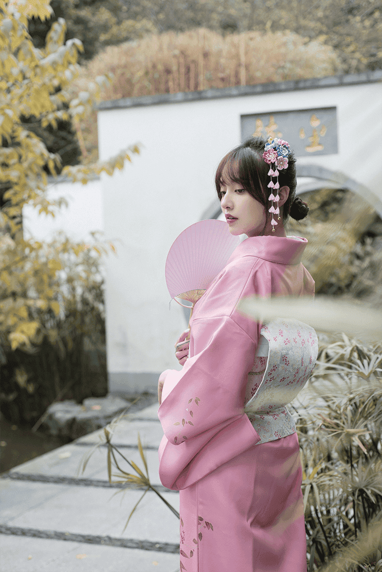 Side profile of a pink Japanese kimono yukata with floral accents, paired with a fan and traditional hair accessories, in a serene setting.