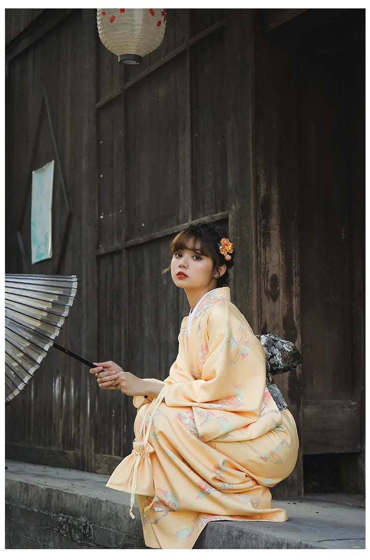 Woman in an orange Japanese kimono-style yukata with floral patterns, sitting near a wooden wall, holding a black umbrella.