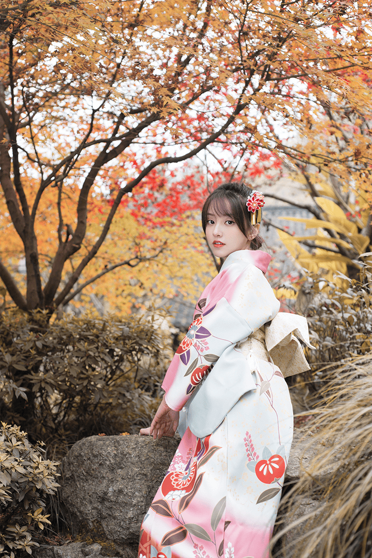 Woman in pink floral kimono posing in a garden with vibrant autumn foliage.