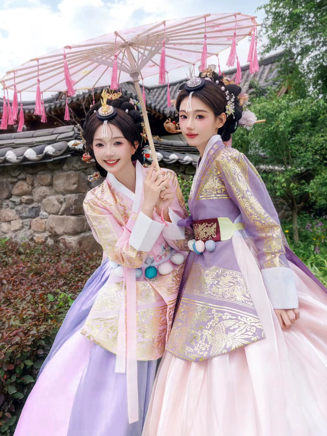 Two women in pink and purple Hanbok holding a traditional parasol in a serene outdoor setting.