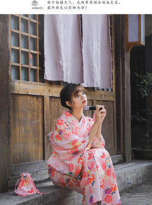 Seated woman in a pink cherry blossom kimono, holding a fan and a matching floral pouch, near a traditional wooden building.