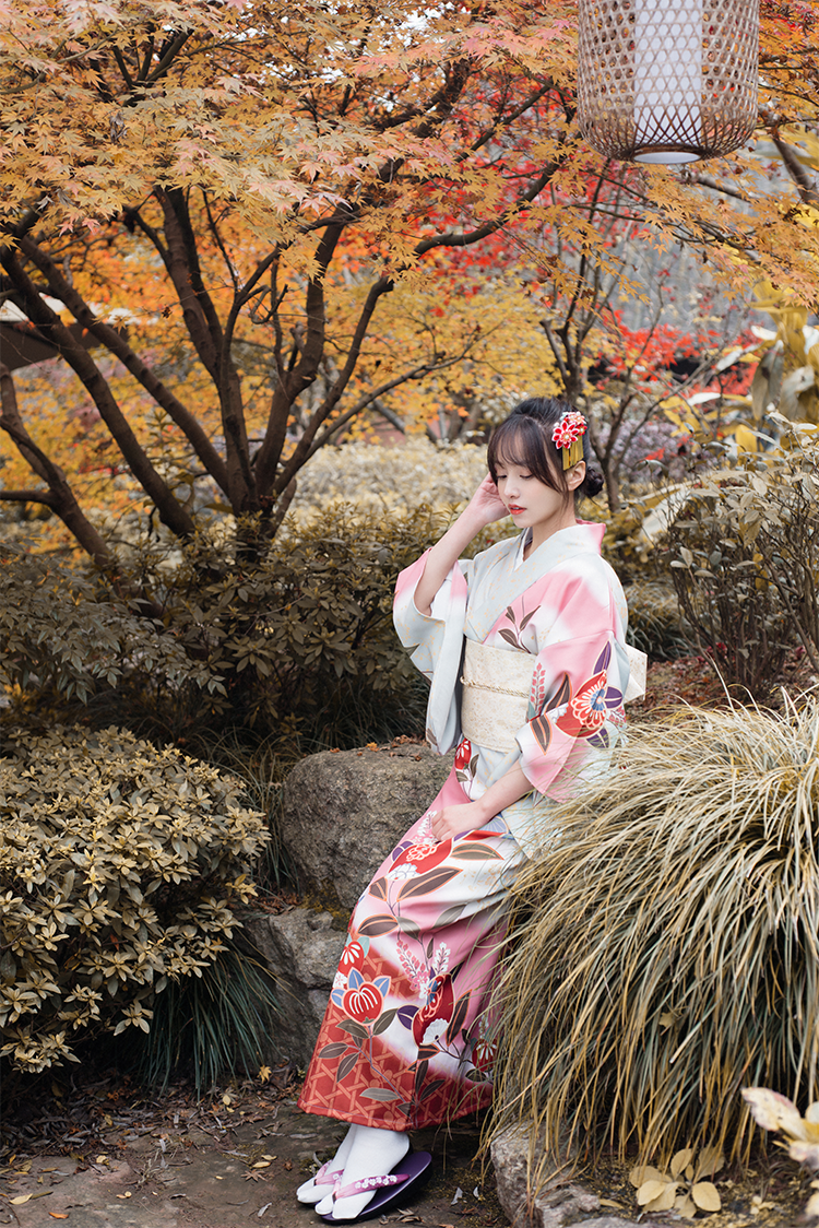 Woman in pink floral yukata sitting in a garden with autumn-colored trees.