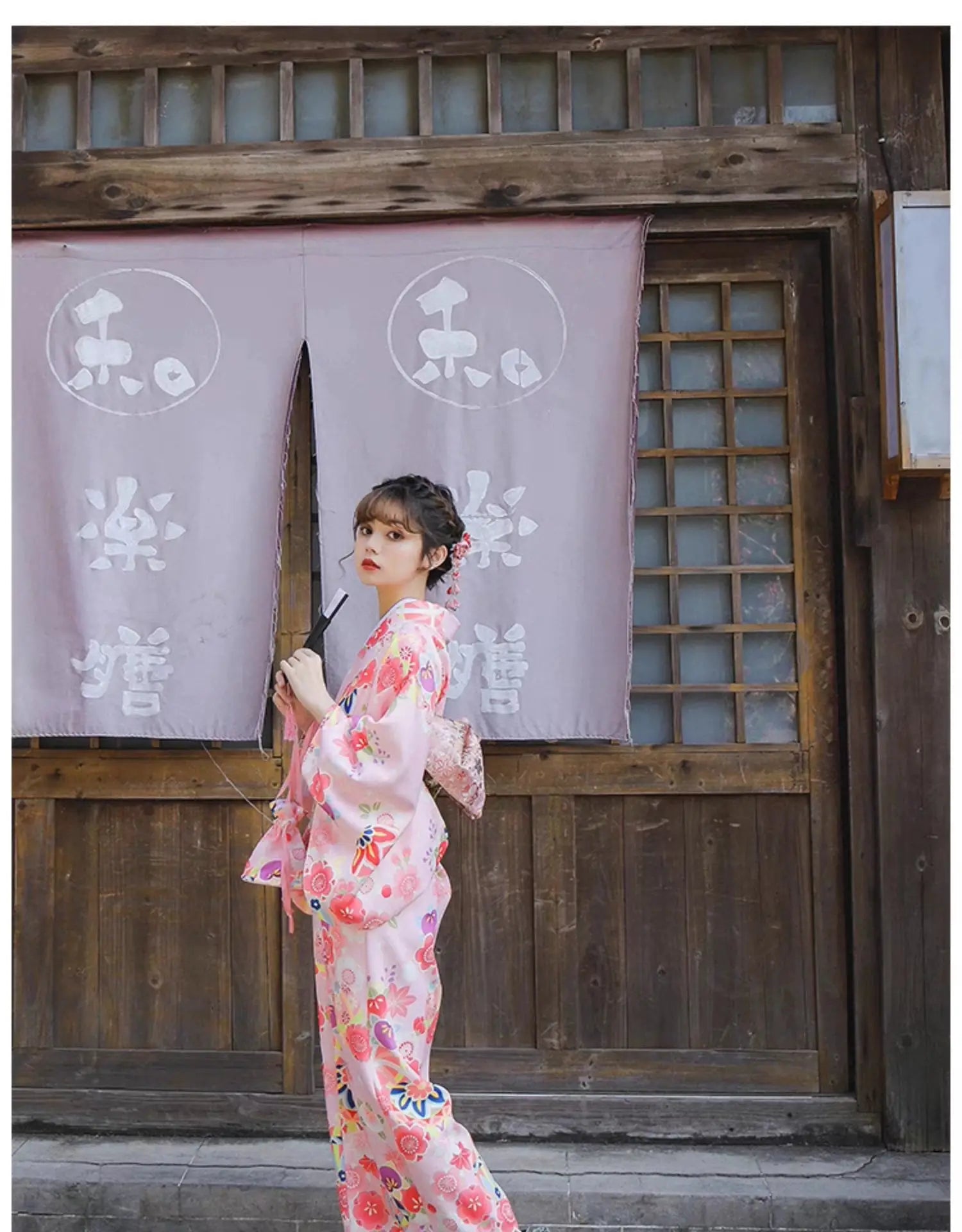 Woman in a pink floral kimono holding a fan, posing in front of a traditional Japanese wooden building with purple curtains.