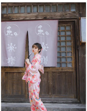 Woman in a pink floral kimono holding a fan, posing in front of a traditional Japanese wooden building with purple curtains.