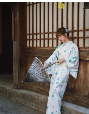 Graceful pose of a woman in a white and blue floral Yukata Kimono Dress, holding a traditional Japanese fan against a wooden backdrop.