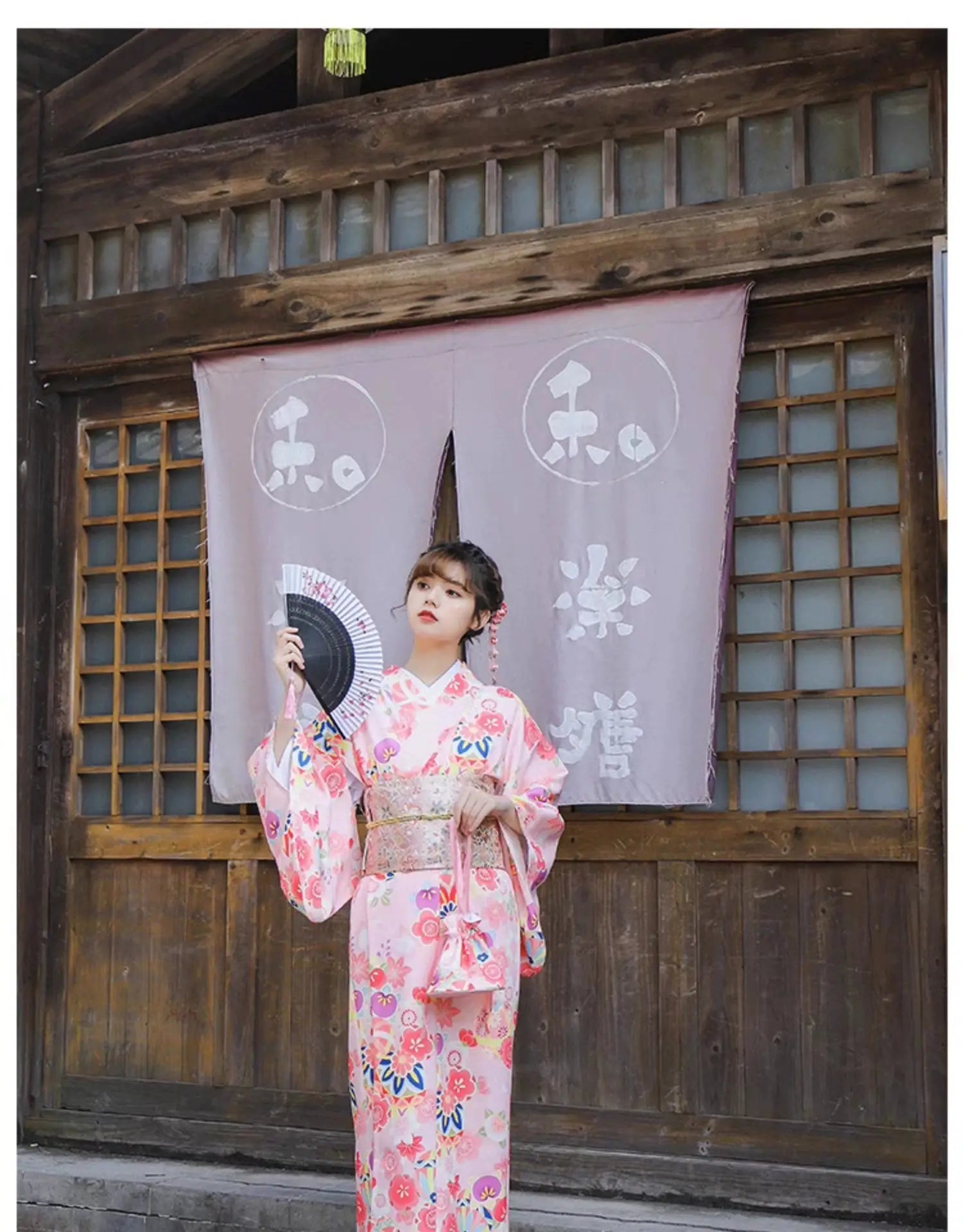 Woman in a pink cherry blossom kimono holding a fan, posing in front of a traditional wooden structure with Japanese curtains.