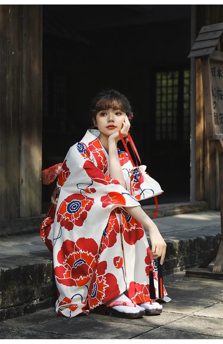 Woman in a white and red floral Japanese kimono dress, holding a traditional accessory, styled in a cultural outdoor setting.