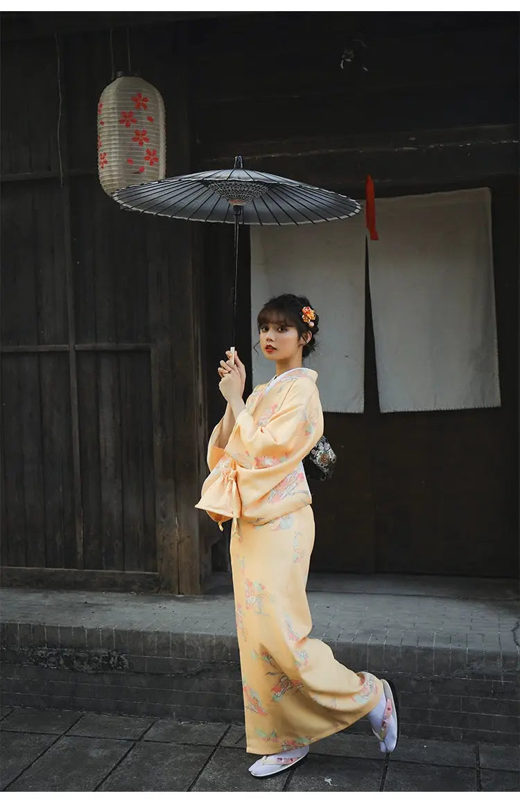 Woman in a formal orange Japanese kimono-style yukata with floral prints, holding a black umbrella under traditional lanterns.