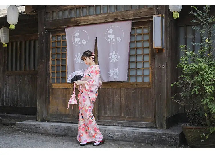 Woman in a pink cherry blossom kimono holding a fan and a floral pouch, standing near a traditional Japanese wooden building.