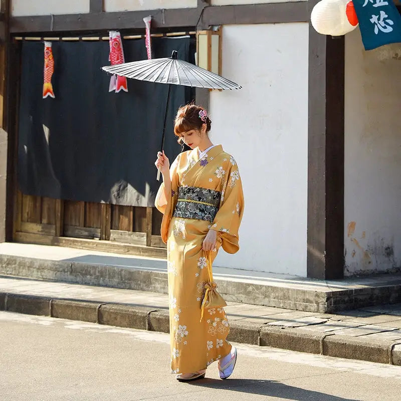 Woman in an orange floral kimono yukata holding a traditional Japanese umbrella, standing in front of a wooden building with lanterns.