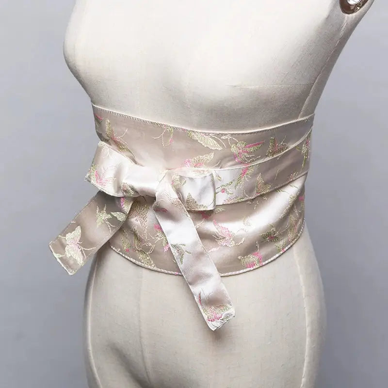 White brocade kimono belt seal with pink and green floral embroidery, tied in a bow on a mannequin for a traditional Japanese look.