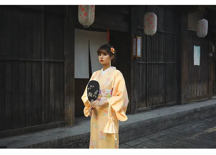 Woman in a formal orange Japanese kimono-style yukata with floral prints, holding a decorative fan in a traditional wooden setting.