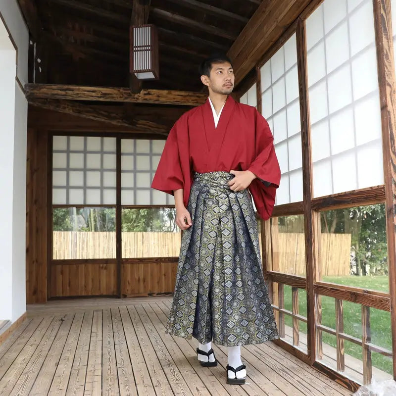 Full-body view of a man in a Japanese formal samurai costume featuring a red kimono and brocade skirt, standing on a wooden deck.