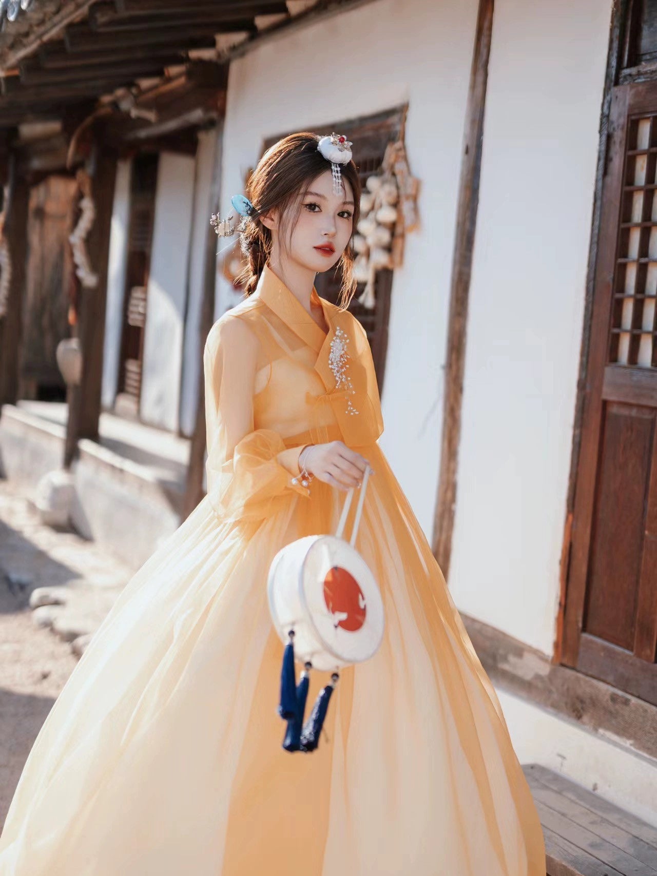 Traditional Korean Hanbok dress in vibrant orange, worn by a woman walking through a historic village, carrying a round embroidered handbag.