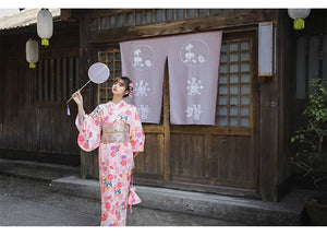 Woman in a pink floral kimono holding a fan, posing near a traditional Japanese wooden building with lanterns and purple curtains.