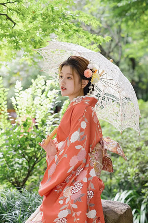 Side profile of a woman in a retro orange kimono with chrysanthemum patterns, holding a lace parasol in a natural setting.