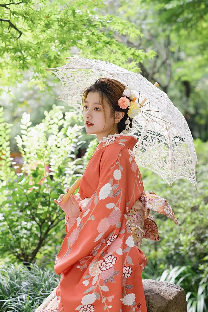 Side profile of a woman in a retro orange kimono with chrysanthemum patterns, holding a lace parasol in a natural setting.