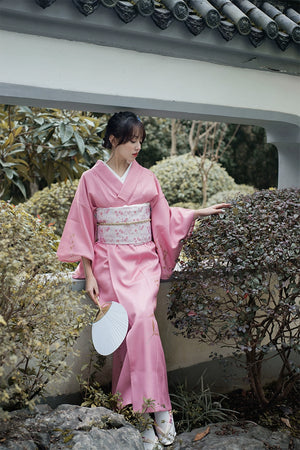 Pink Japanese kimono yukata with floral accents, styled with a white obi and fan, worn in a tranquil garden environment.