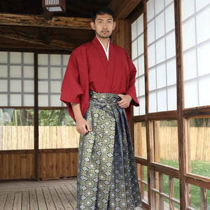 Man wearing a Japanese traditional kimono with a red top and patterned brocade skirt, standing in a wooden room with shoji screens.