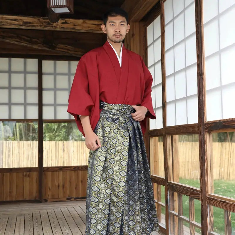 Man wearing a Japanese traditional kimono with a red top and patterned brocade skirt, standing in a wooden room with shoji screens.