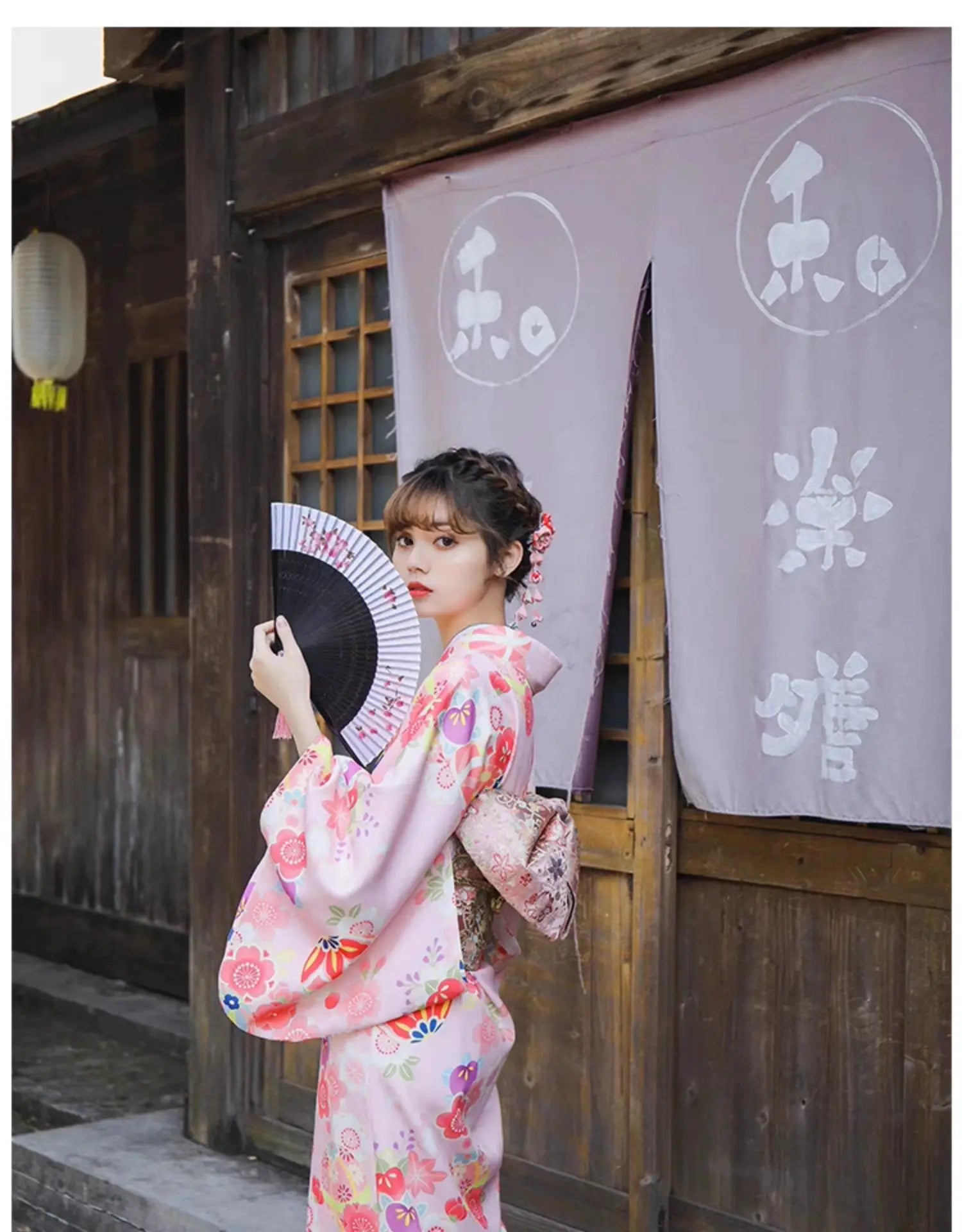 Woman in a pink floral kimono holding a fan, posing with a traditional Japanese wooden building as the backdrop.