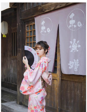 Woman in a pink floral kimono holding a fan, posing with a traditional Japanese wooden building as the backdrop.