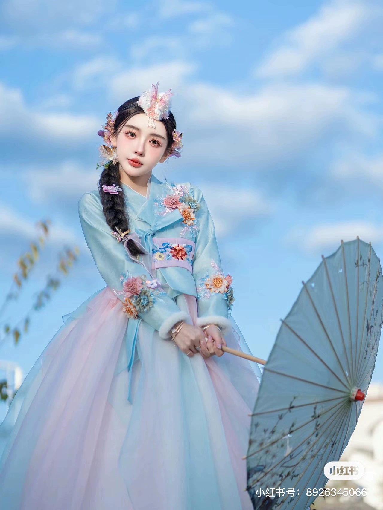 Graceful blue Hanbok with 3D floral design, complemented by a pastel parasol and clear sky.