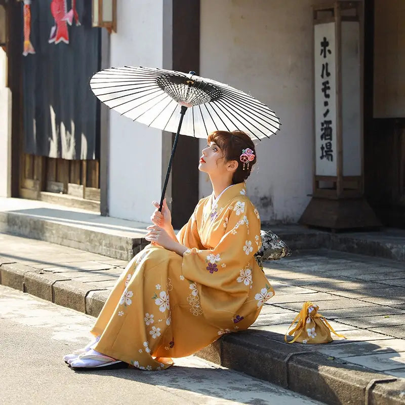 Woman in an orange floral kimono yukata sitting on a stone pavement, holding a traditional umbrella under soft daylight.