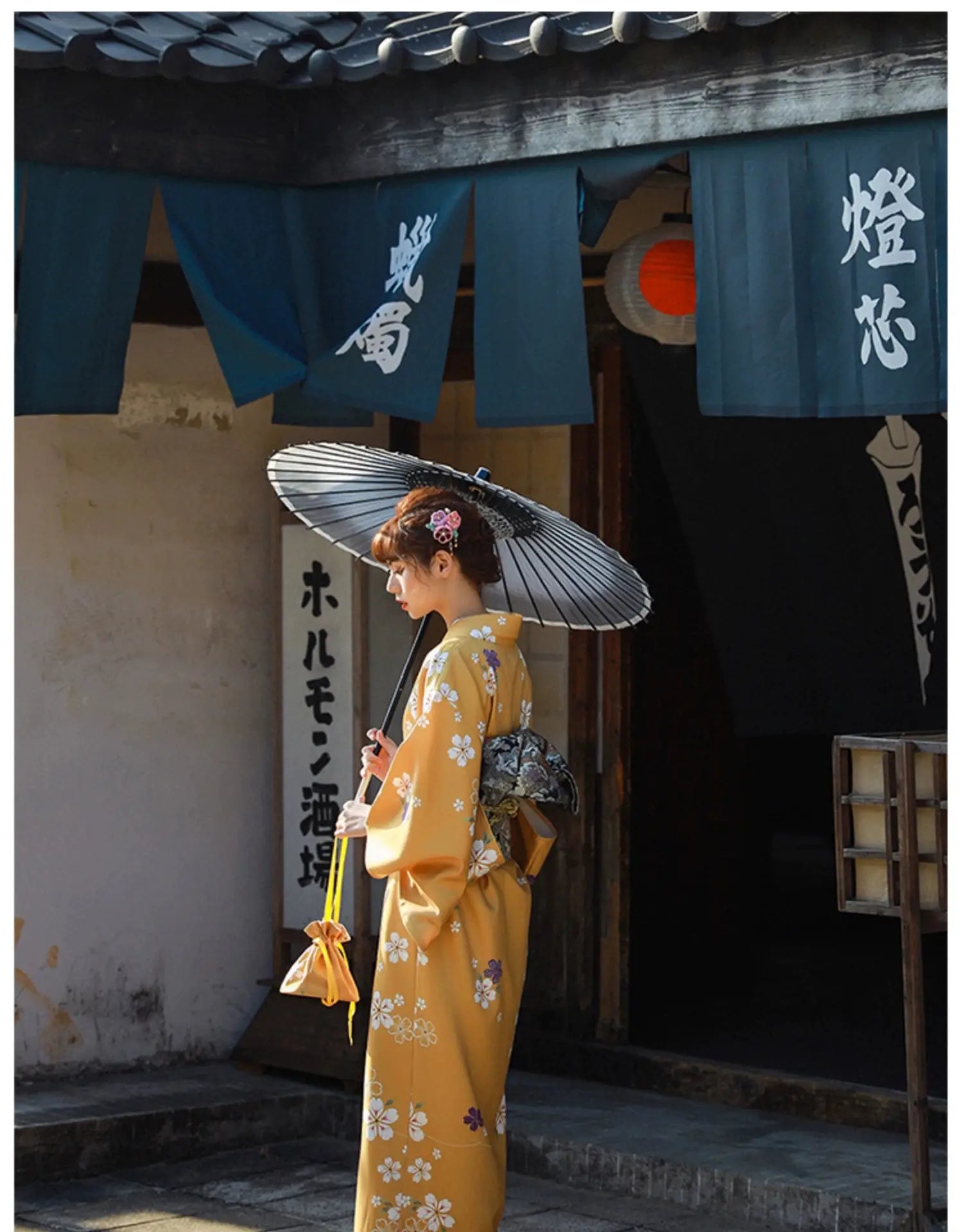 Back view of a woman in an orange floral kimono yukata holding a traditional umbrella, standing near a Japanese building with blue banners.