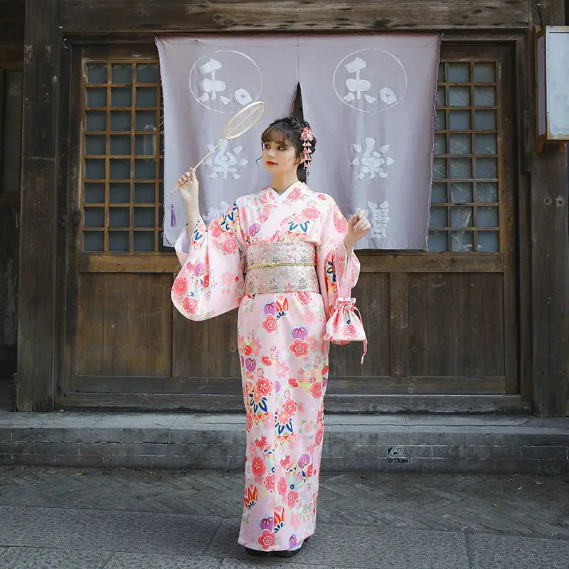 Japanese woman wearing a pink cherry blossom kimono with floral patterns, holding a decorative fan in front of a traditional wooden building.