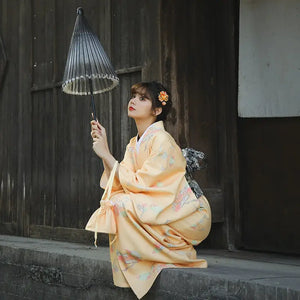 Woman in an orange Japanese kimono-style yukata with floral patterns, holding a traditional umbrella, sitting near a wooden wall.