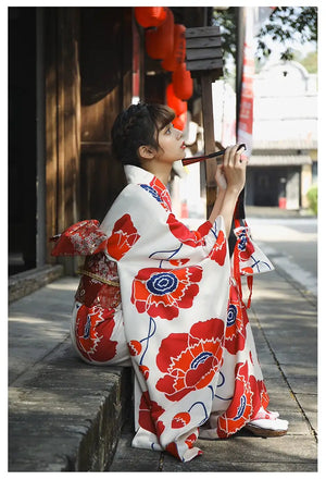 Side profile of a woman in a white and red floral Japanese kimono dress, holding a traditional pipe in a serene outdoor setting.