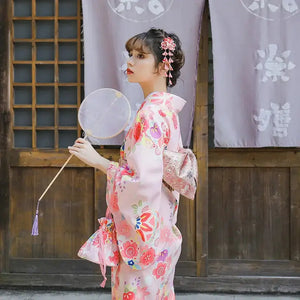 Close-up of a woman in a pink cherry blossom kimono holding a round fan, showcasing intricate floral patterns and traditional Japanese accessories.