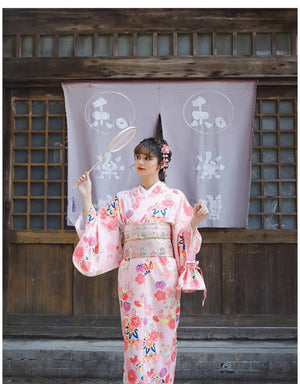 Woman in a pink cherry blossom kimono holding a round fan, standing in front of a traditional Japanese wooden structure.