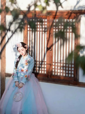 Model in blue Hanbok with floral accents, leaning against a traditional wooden door.