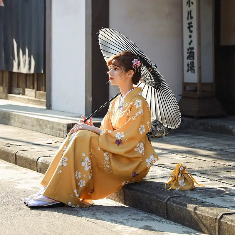 Seated woman in an orange floral kimono yukata holding a black and white umbrella, with a matching bag placed beside her on a stone pavement.