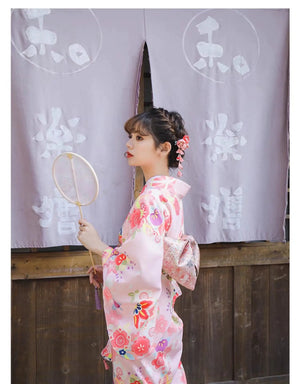 Woman in a pink floral kimono holding a round fan, posing in front of a traditional Japanese wooden building with purple curtains.