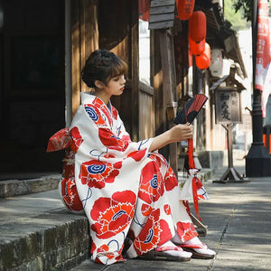 Elegant white kimono dress with red floral designs, worn by a woman seated outdoors, holding a traditional Japanese fan.