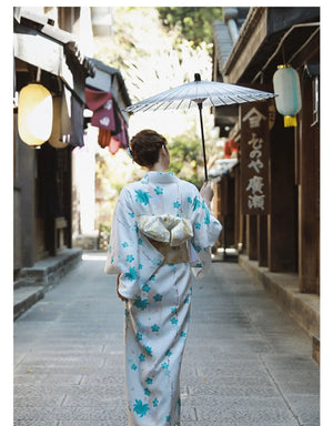 Back view of a woman in a white and blue floral Yukata Kimono Dress with a gold obi, walking under a parasol in a traditional Japanese setting.