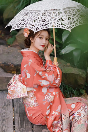 Close-up of a woman in a chrysanthemum-patterned orange kimono holding a lace parasol, styled for photography and travel.