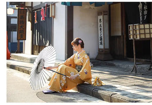 Woman in an orange floral kimono yukata sitting on a stone pavement, holding a traditional umbrella with a serene Japanese street backdrop.