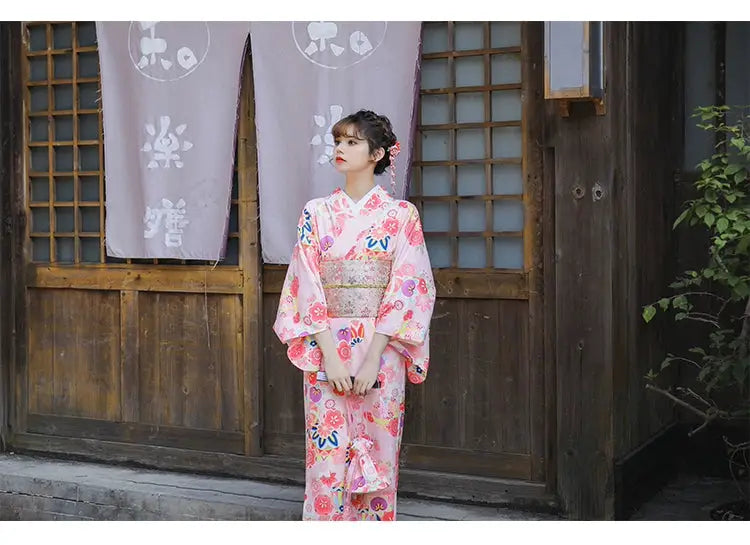 Woman in a pink cherry blossom kimono posing in front of a traditional Japanese wooden building, showcasing intricate floral patterns.