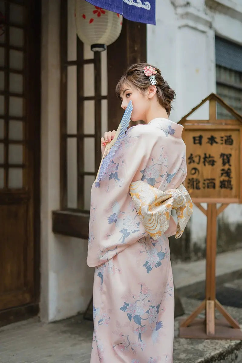 Back view of a woman in a pink yukata kimono with crane motifs, featuring a gold obi, standing near traditional Japanese decor.