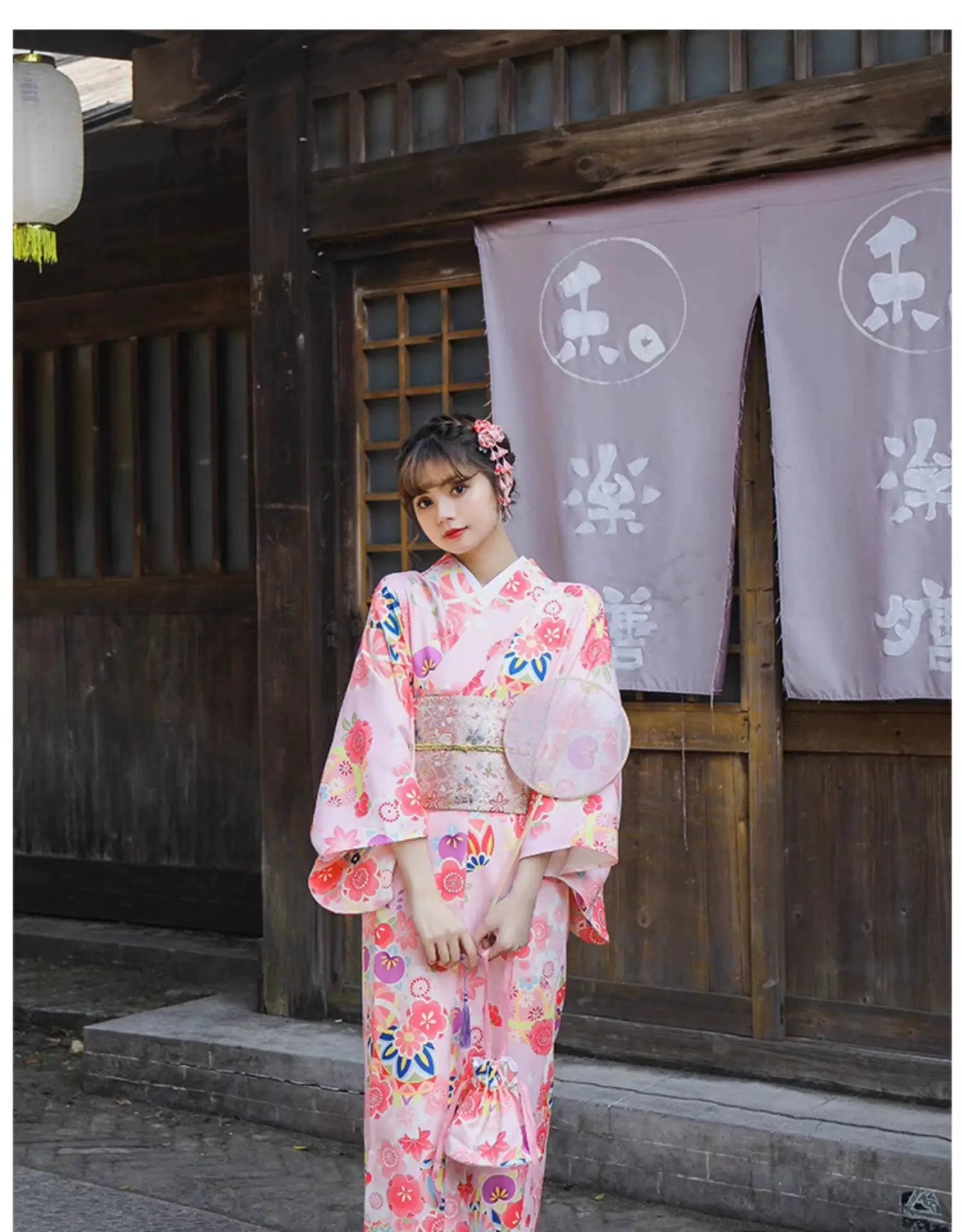 Woman in a pink cherry blossom kimono holding a round fan, standing in front of a traditional Japanese wooden building.