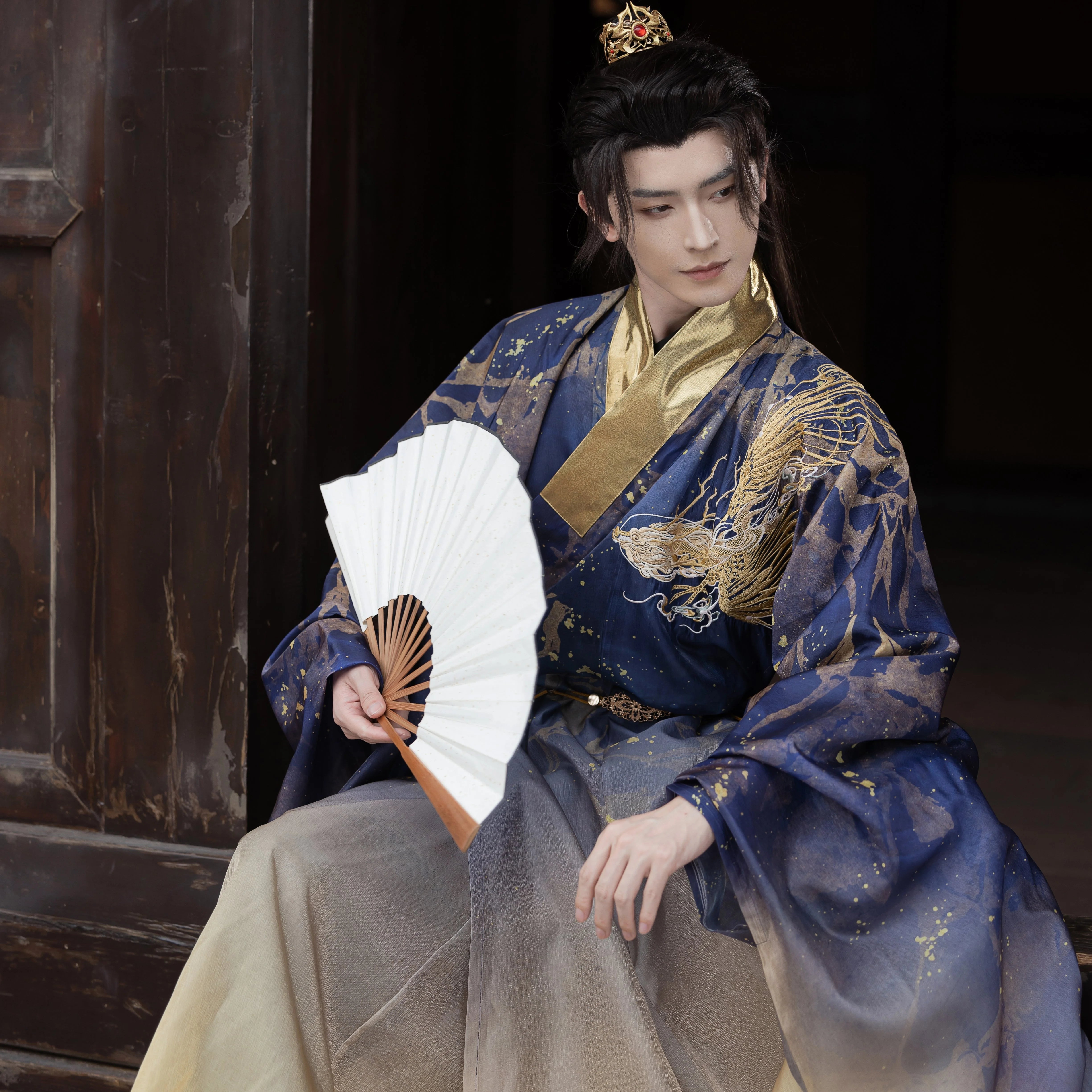 Model seated in a Dragon Roar Hanfu Daoist Robe, holding a white fan, showcasing the Ming blue fabric with gold embroidery details.