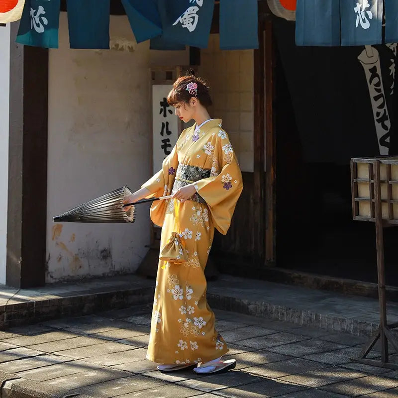 Woman in an orange floral kimono yukata walking with a folded umbrella, set against a traditional Japanese building with blue banners.