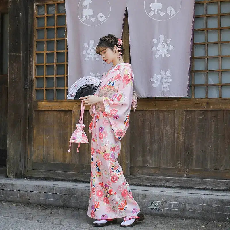 Side view of a woman in a pink floral Japanese kimono holding a fan, standing near a traditional wooden structure with purple curtains.