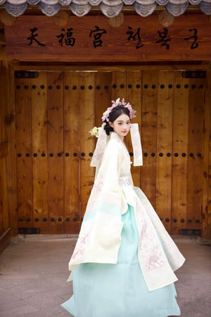 Traditional Korean wedding dress in soft yellow by Muzhi, adorned with intricate embroidery and pastel accents for an elegant bridal ensemble.