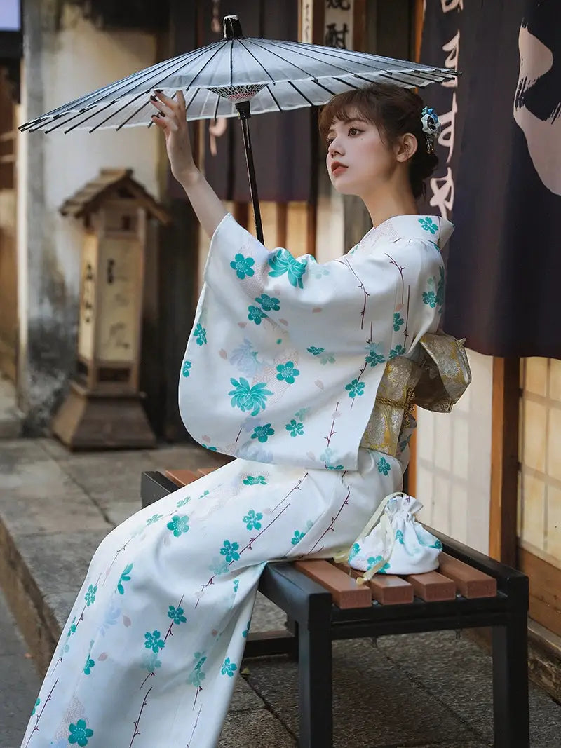 Seated woman in a white and blue floral Yukata Kimono Dress, holding a traditional Japanese parasol, showcasing cultural elegance.